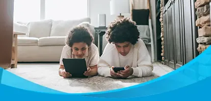 Two children lie on a living room floor, each focused on a tablet and smartphone.